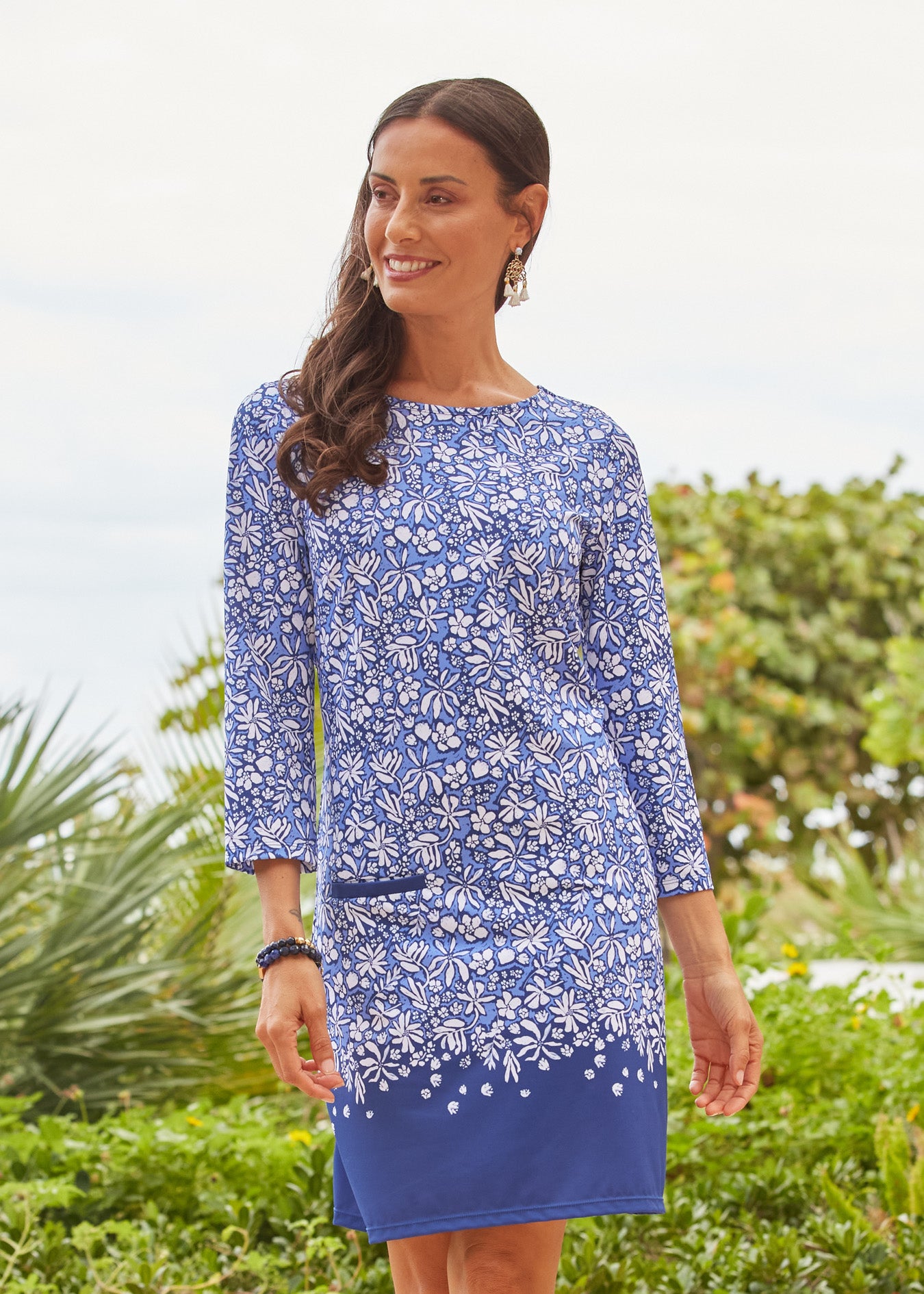 A brunette woman walking in front of palms wearing the sun protective Seaside Floral Cabana Shift Dress from Cabana Life.