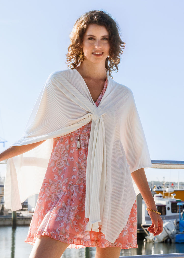 A woman standing outdoors by a marina wearing a flowing white wrap tied at the front over a pink floral dress.