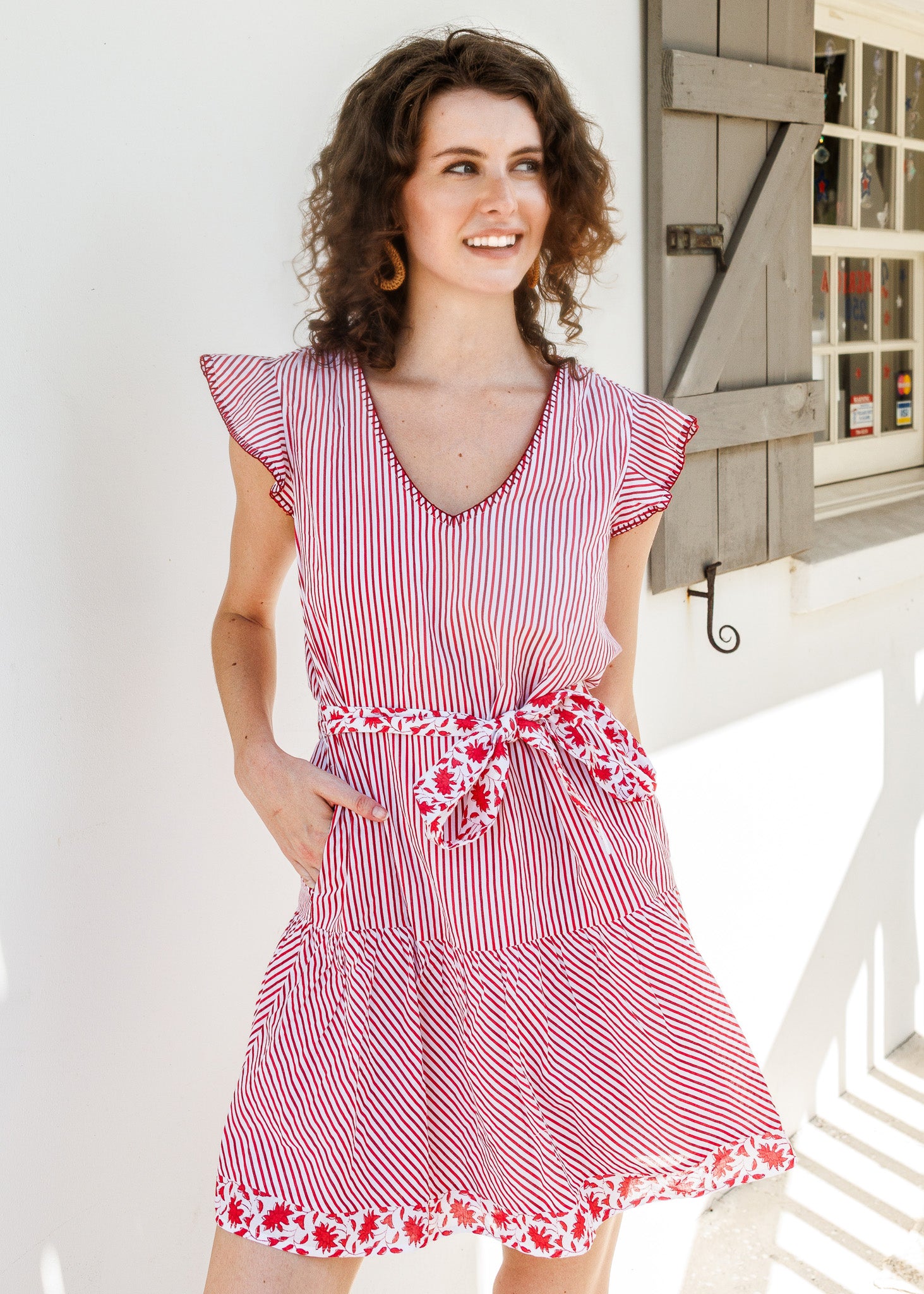 A woman wearing a red and white vertically striped tiered dress with ruffled cap sleeves, a matching tie belt, and a floral patterned hem, standing outdoors against a white wall.