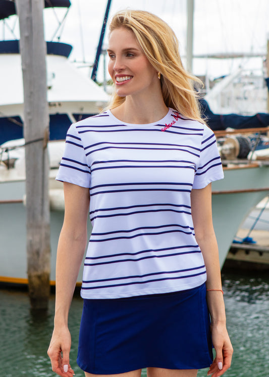 Woman wearing a striped shirt and blue skirt standing by a marina with boats in the background.