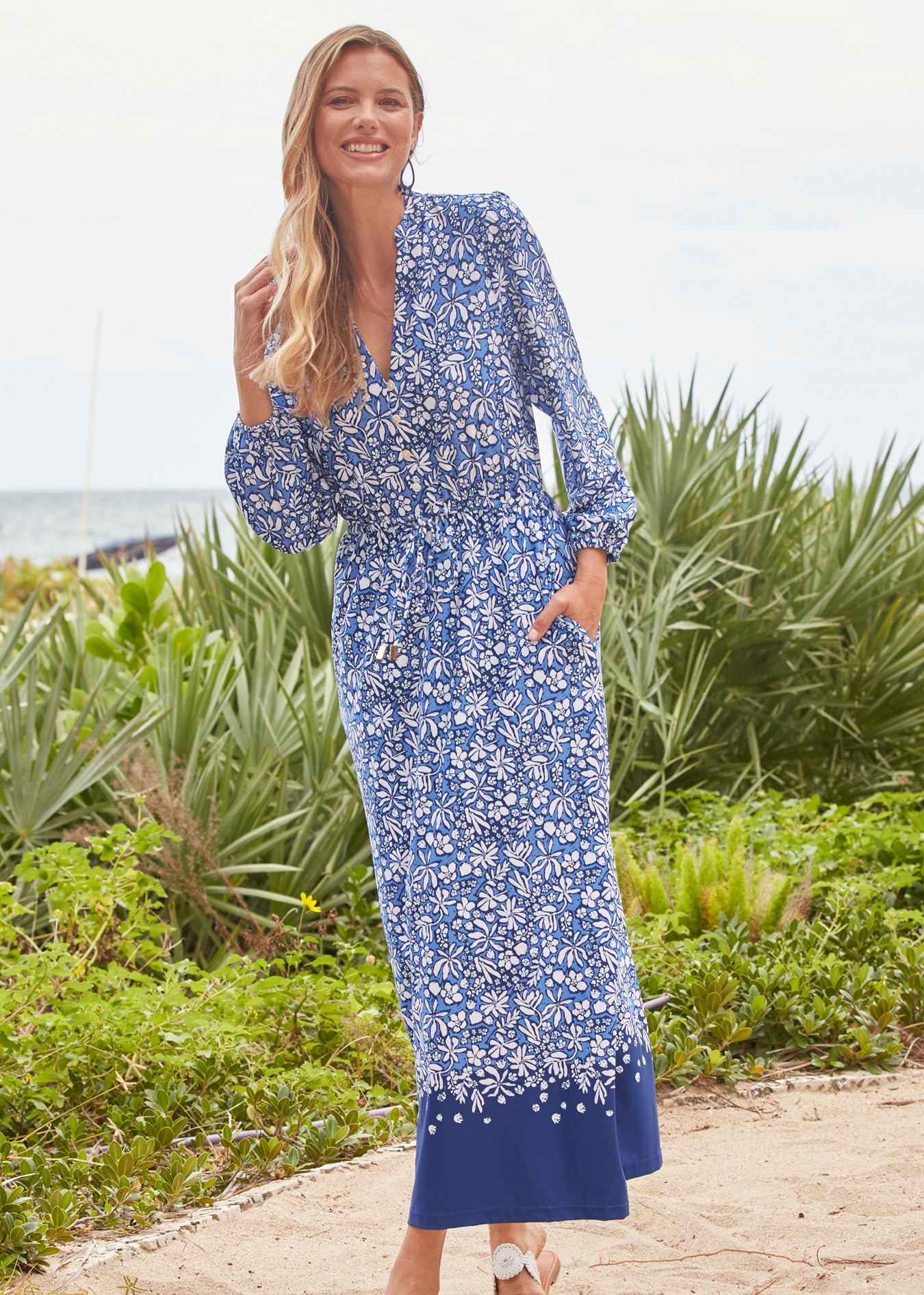 Woman wearing a blue floral dress standing outdoors with greenery in the background