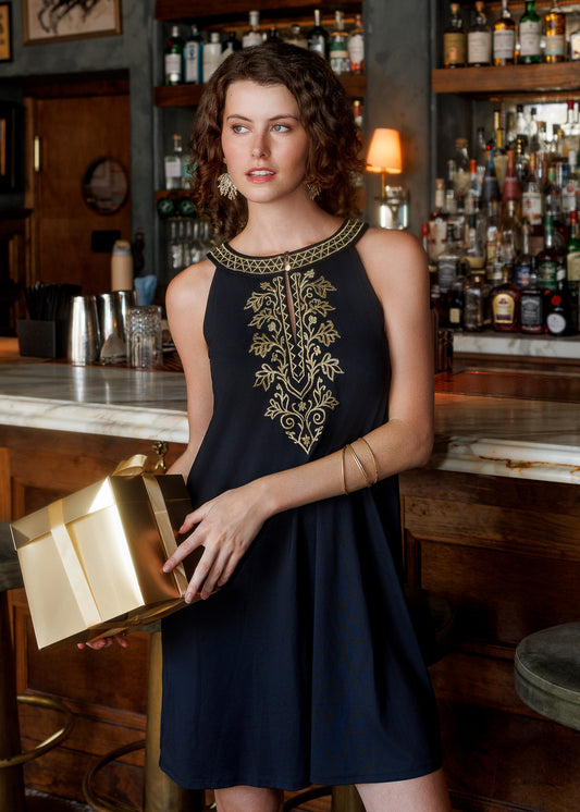 A woman wearing a sleeveless blacl dress with gold embroidery on the chest, holding a metallic gold gift box, posed in front of a bar with liquor bottles in the background.