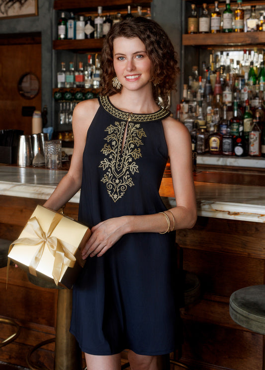 A woman smiling wearing a sleeveless blacl dress with gold embroidery on the chest, holding a metallic gold gift box, posed in front of a bar with liquor bottles in the background.