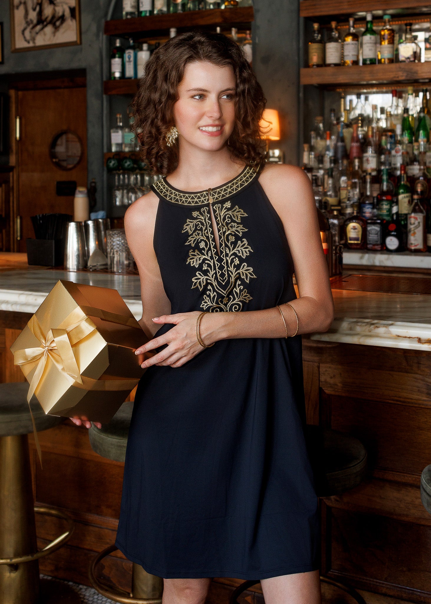 A woman wearing a sleeveless blacl dress with gold embroidery on the chest, holding a metallic gold gift box, posed in front of a bar with liquor bottles in the background.
