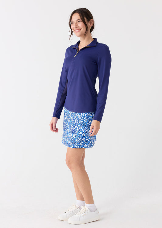 The side of a dark haired woman wearing the Cabana Life sun protective Navy 1/4 Zip Sport Top and Seaside Floral 17" Skort with white crew socks and sneakers.