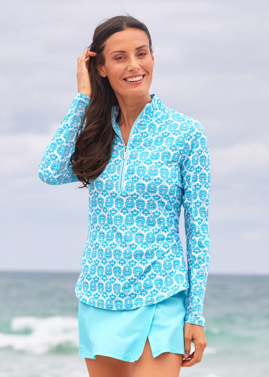 A brunette woman wearing the Cabana Life sun protective Rosemary Beach Performance 1/4 Zip Sport Top with mesh underarms, paired with the Aqua Classic Swim Skirt on the beach.