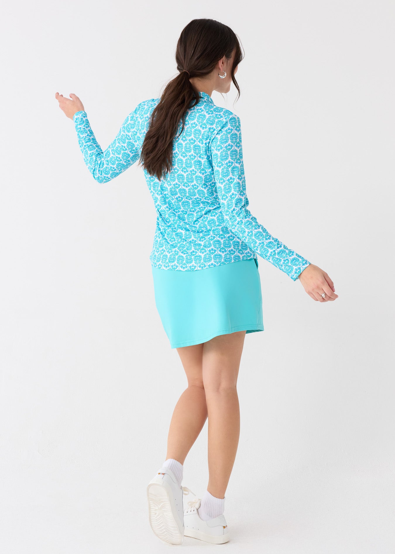 The back of a brunette woman wearing the Cabana Life sun protective Rosemary Beach Performance 1/4 Zip Sport Top and Aqua 17" Skort with white sneakers.