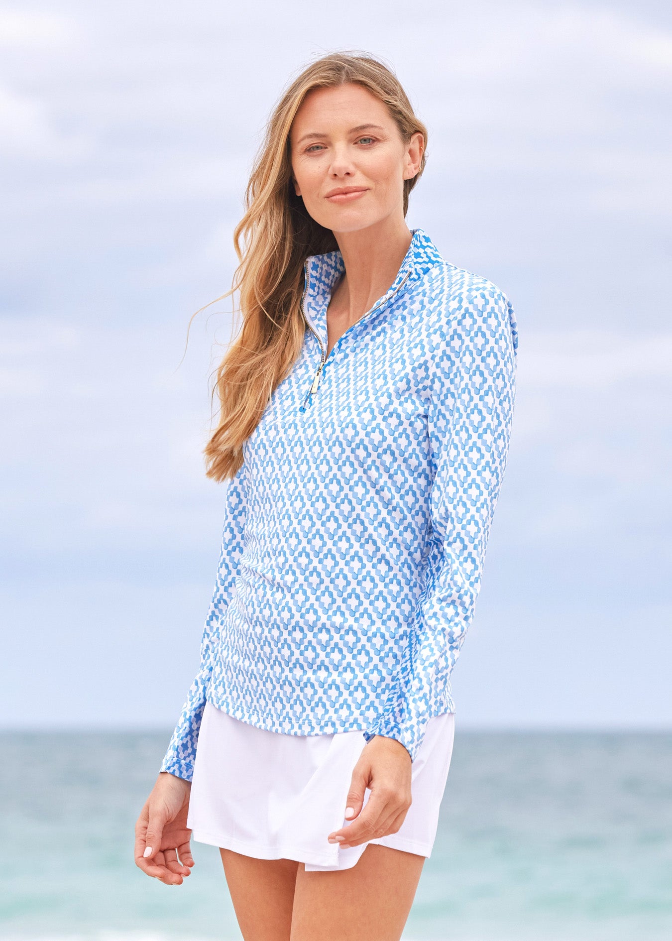 A blonde woman to the side wearing the Cabana Life sun protective Seaside 1/4 Zip Sport Top and White Classic Swim Skirt on the beach.