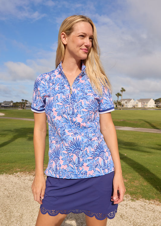 A blonde woman wearing the Cabana Life sun protective Palm Beach Short Sleeve Collared 1/4 Zip with the Navy 15" Scallop Skort in front of a golf course.