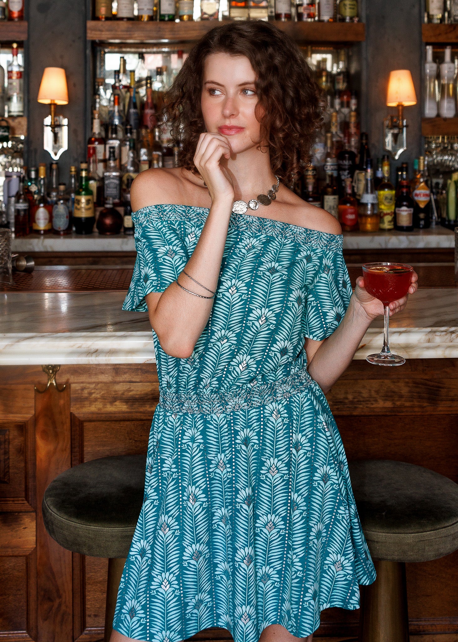 A woman wearing a teal and white palm leaf print off-the-shoulder dress stands in a bar setting, holding a red cocktail glass.