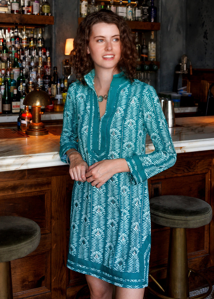 A woman wearing a teal and white patterned long-sleeve tunic dress stands in a bar setting with wooden stools and shelves of bottles in the background.