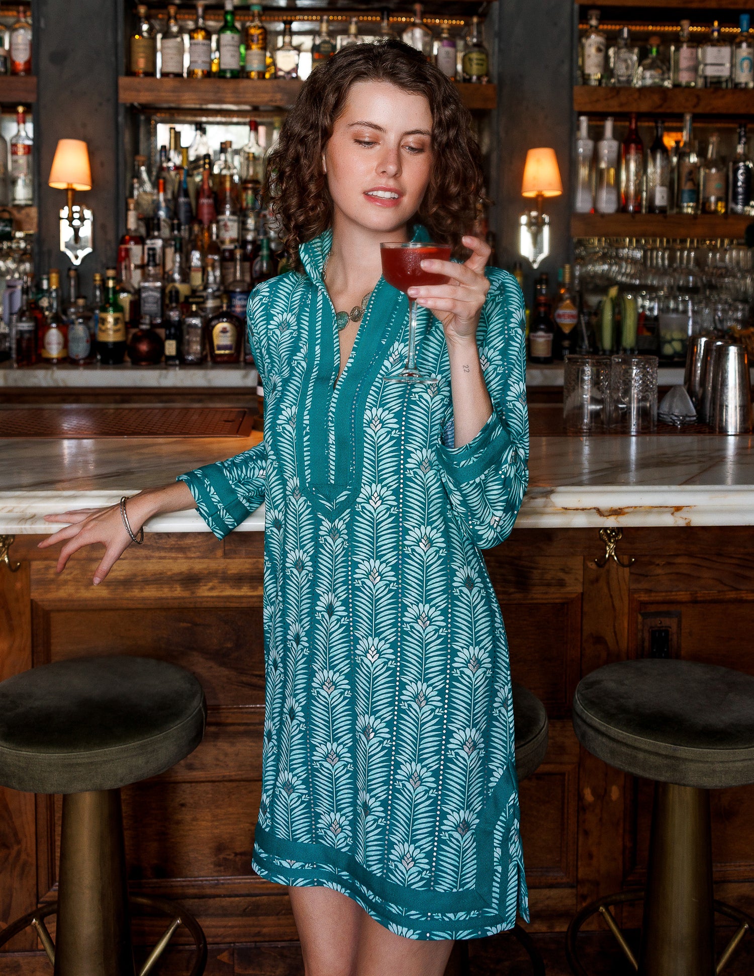A woman wearing a teal and white printed dress stands in a bar setting with wooden stools and shelves of bottles in the background.