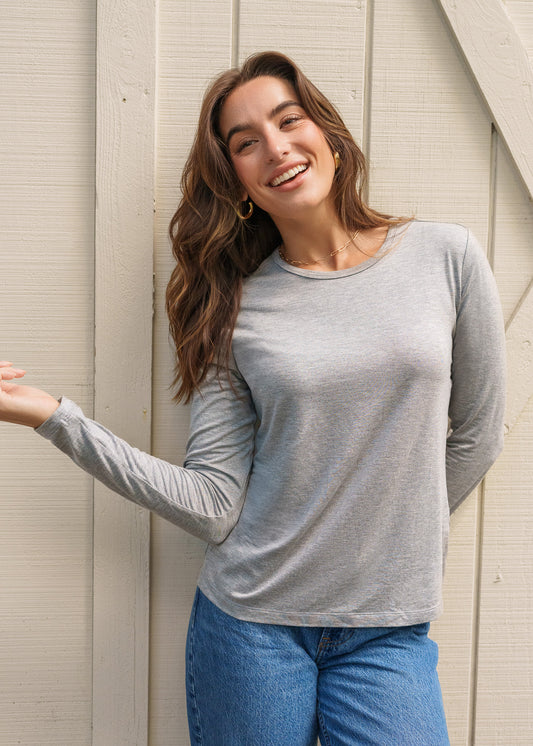 Woman wearing a gray long-sleeve top and blue jeans standing against a light wooden wall.