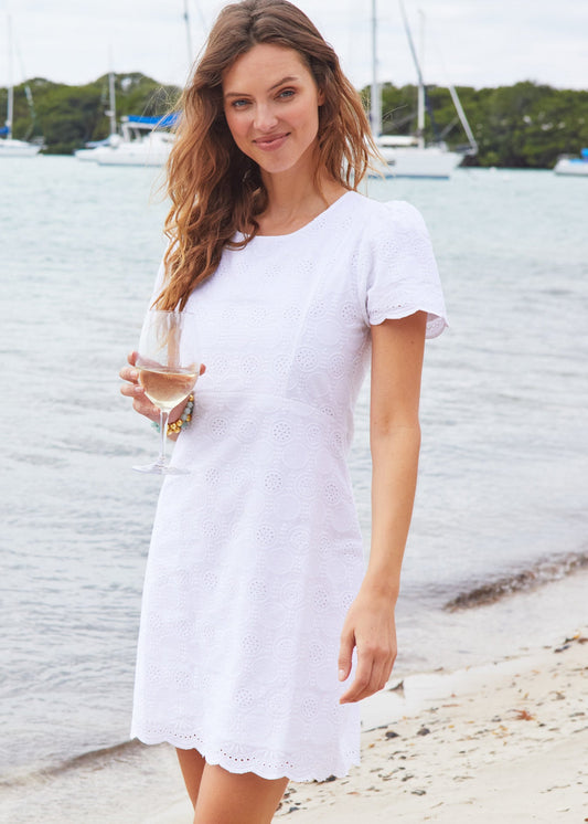 A woman with brown curly hair on the sand wearing the White Eyelet Short Sleeve Shift Dress and holding a wine glass.