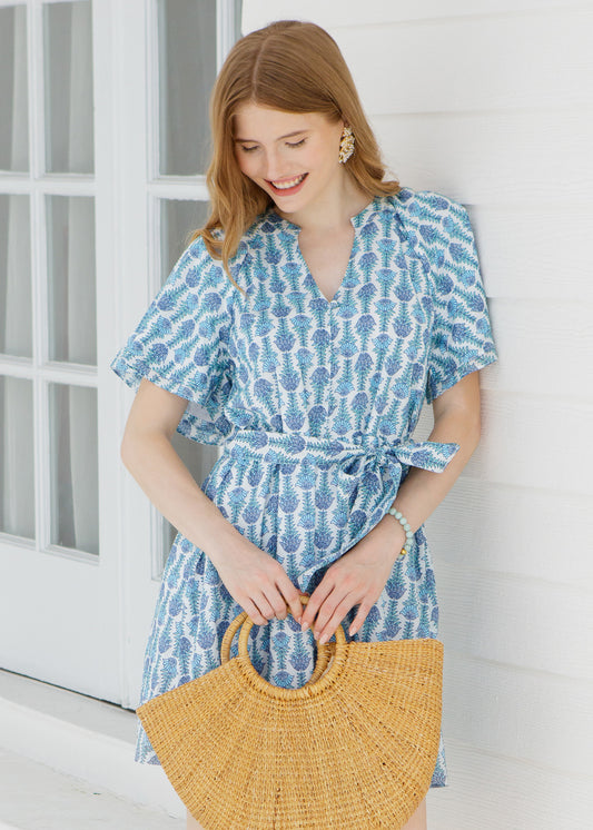 Woman in a blue patterned dress holding a  straw bag against a white background