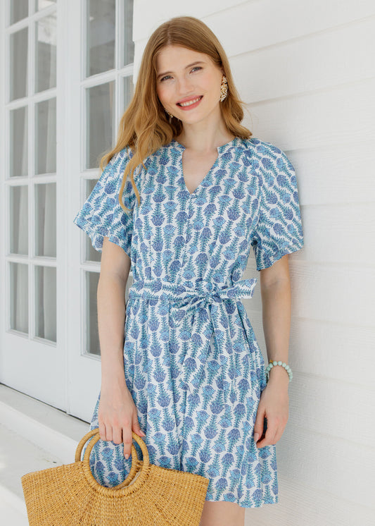 Woman wearing a blue patterned dress holding a straw bag against a white wall.