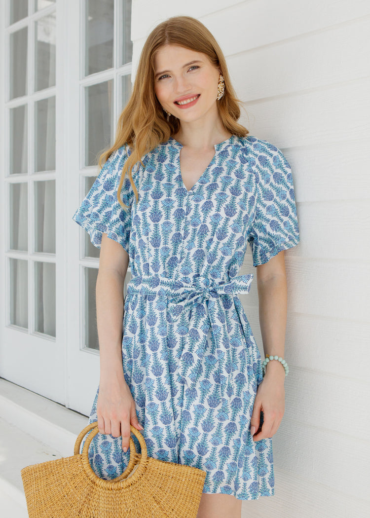 Woman wearing a blue patterned dress holding a straw bag against a white wall.