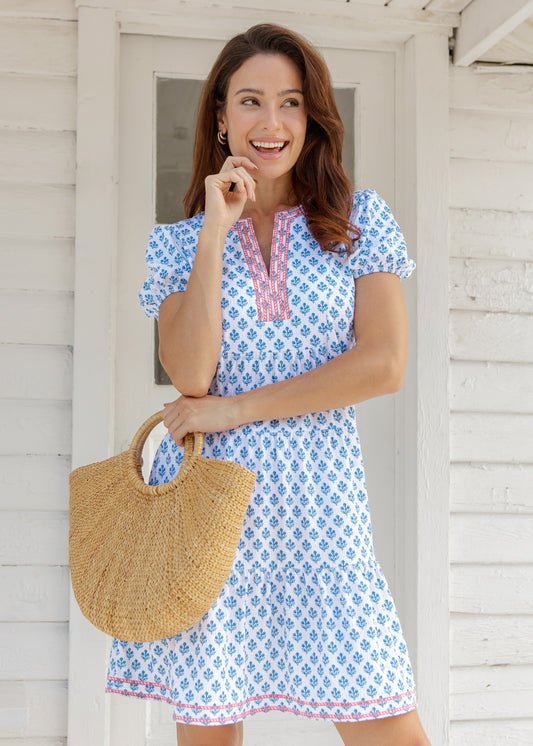 Woman in a blue patterned dress holding a straw bag, standing in front of a white door.