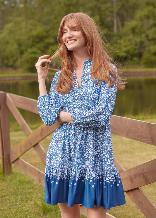 Woman in a blue floral dress standing by a wooden fence with a natural background