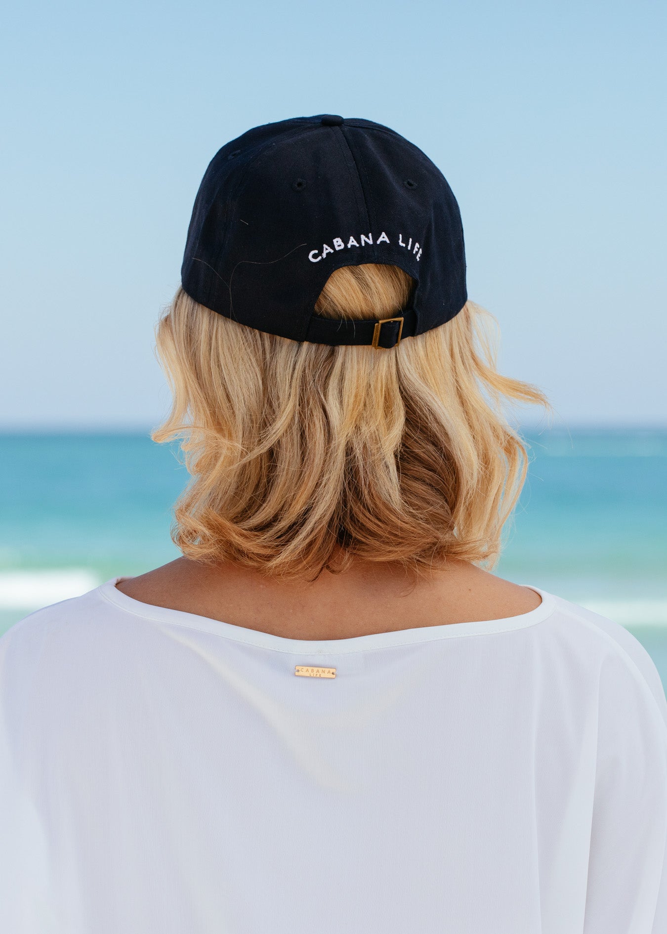 Back of blonde woman wearing Navy Cabana Life Baseball Hat facing beach. 