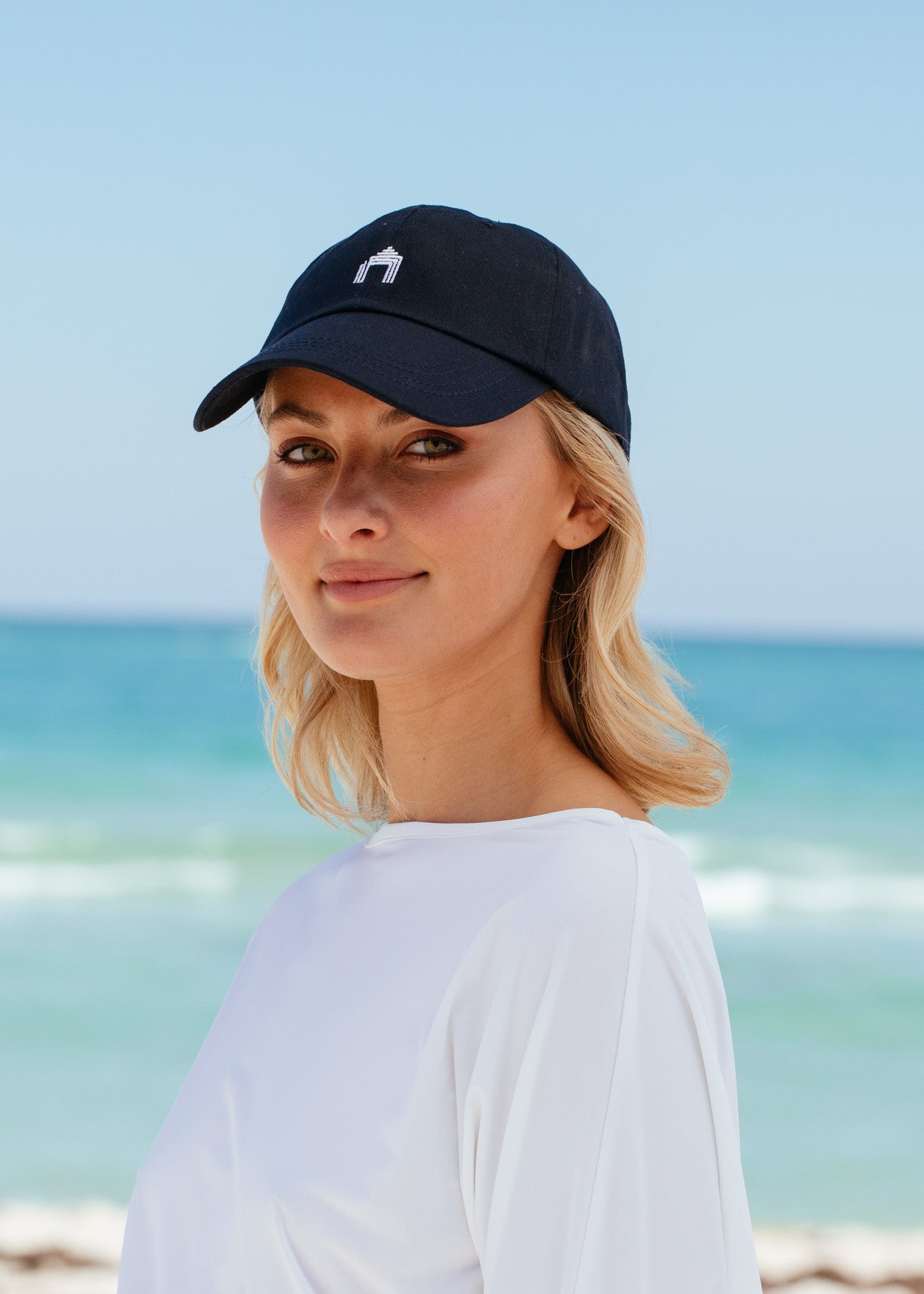 Blonde woman wearing Navy Cabana Life Baseball Hat with white rashguard on beach.