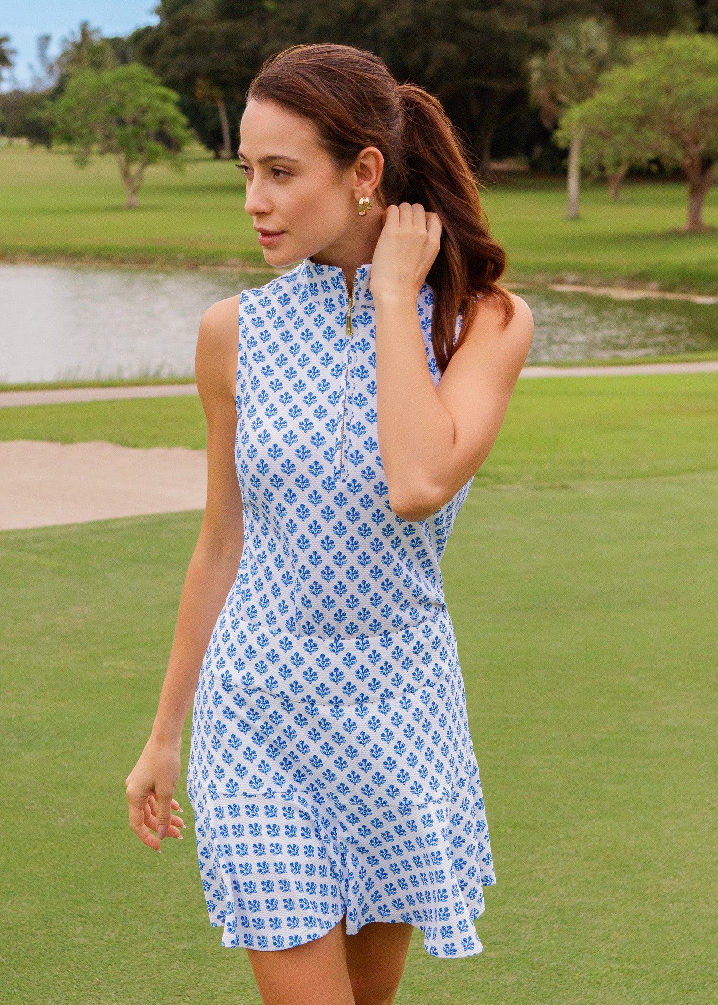 Woman in a blue patterned dress standing on a golf course with trees and water in the background.