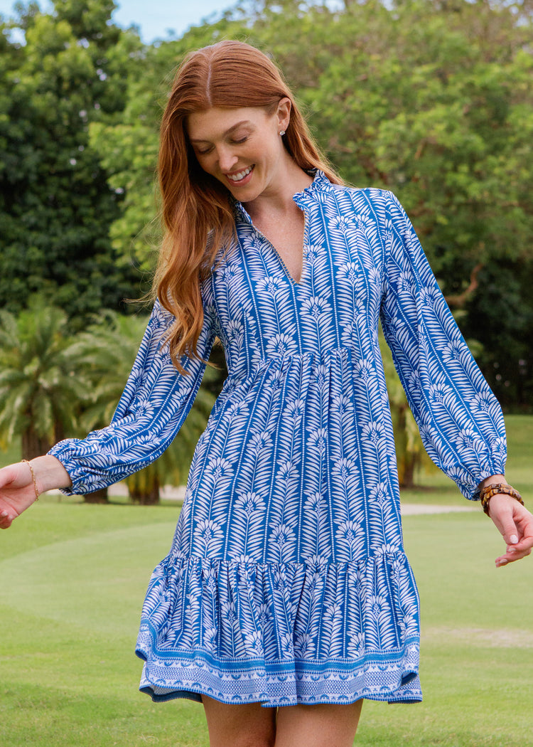 Woman wearing a blue patterned dress standing outdoors with greenery in the background