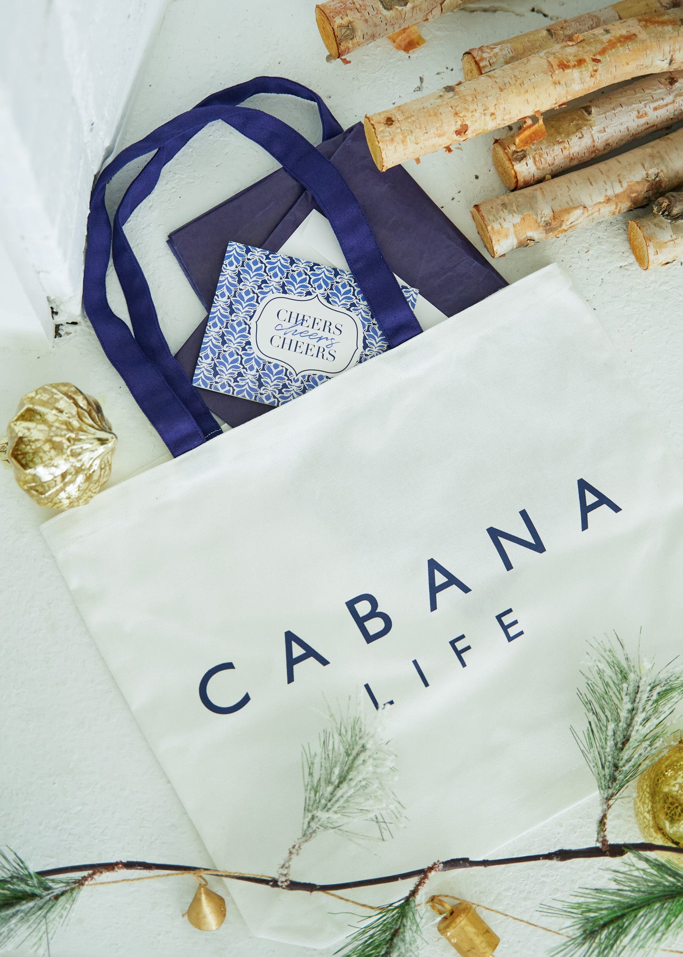 White canvas Cabana Life Tote Bag, navy tissue paper and Cheers greeting card peeking out by ornaments and firewood