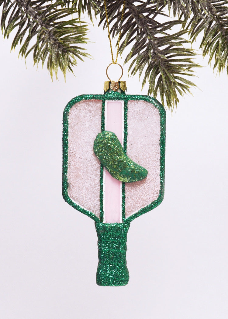Decorative ornament shaped like a pickleball paddle, finished in vibrant shades of pink and green sparkle.  Topped with a glittery green pickle, hanging from a Christmas tree by the metallic gold string.