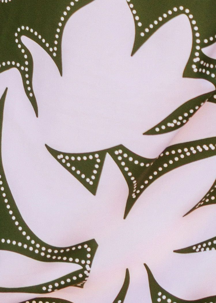 A close-up of a fabric pattern featuring a large pink leaf shape outlined with dark olive green borders decorated with small white dots, set against a dark green background.
