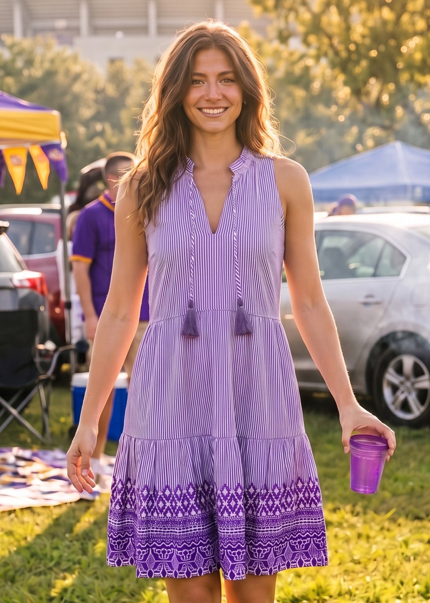 A woman wearing a sleeveless purple and white vertically striped tiered dress with tassel ties and a purple geometric patterned hem, standing outdoors at a sunny event and holding a purple cup.