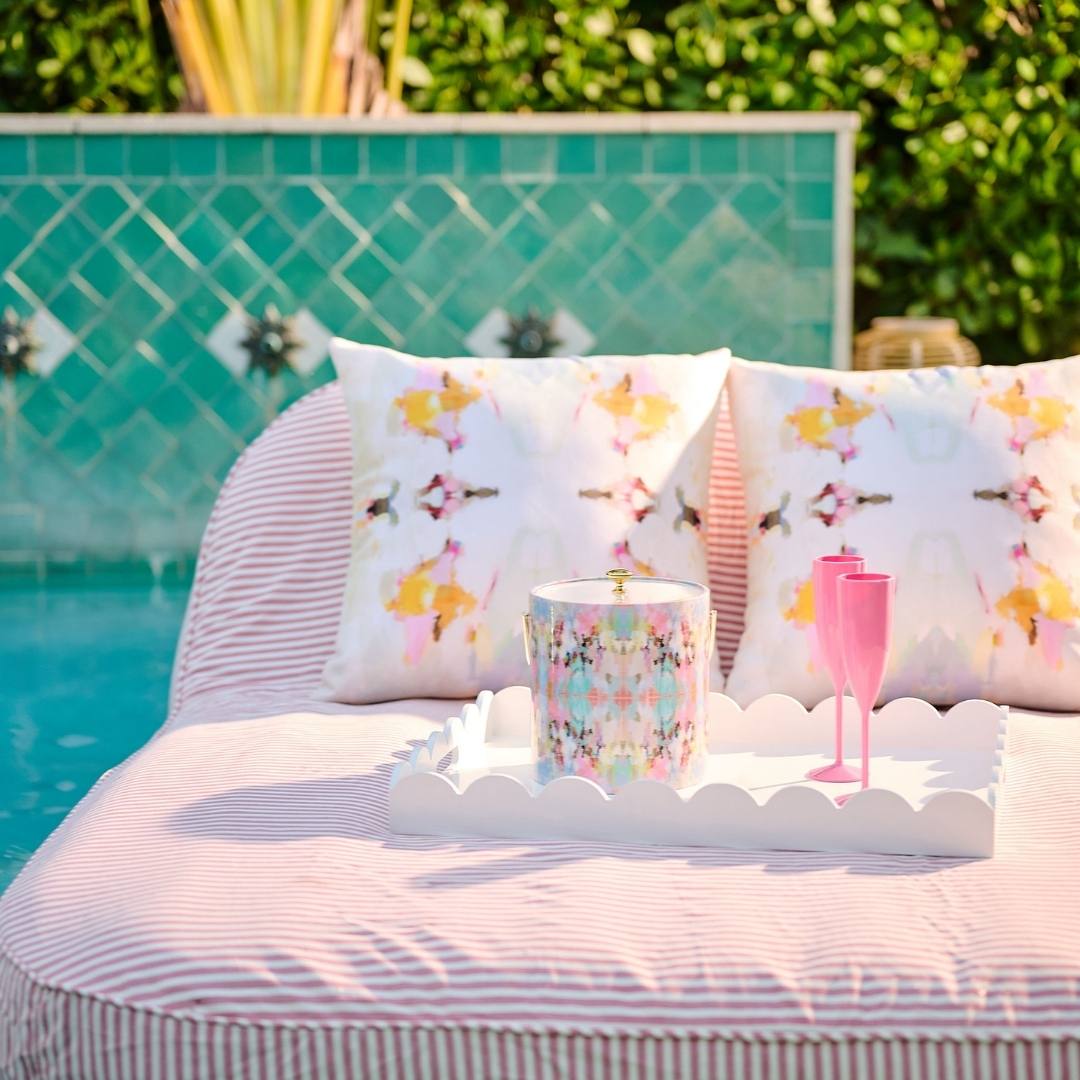 Outdoor setting with a striped sofa, floral cushions, and a tray with a mug and glasses by a pool.