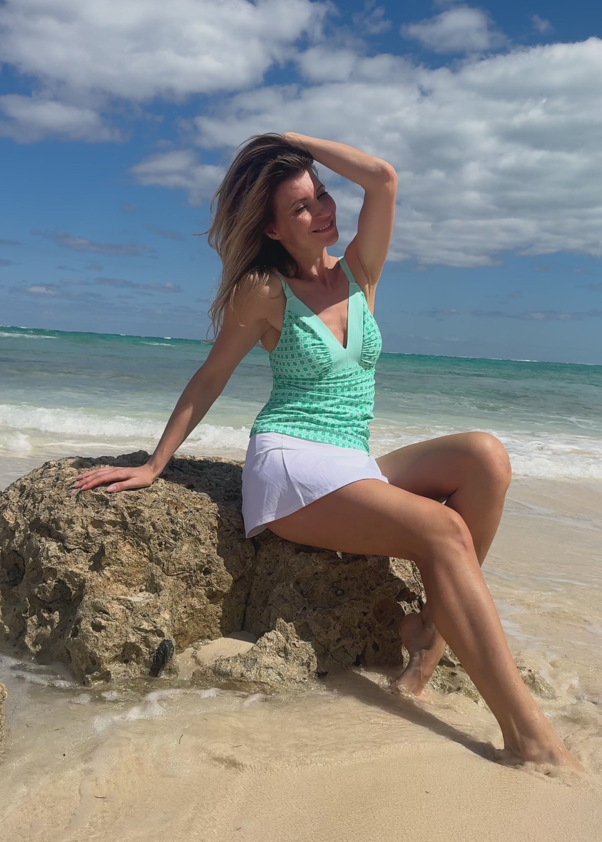 Woman wearing a green patterned tankini top and white skirt on a beach.