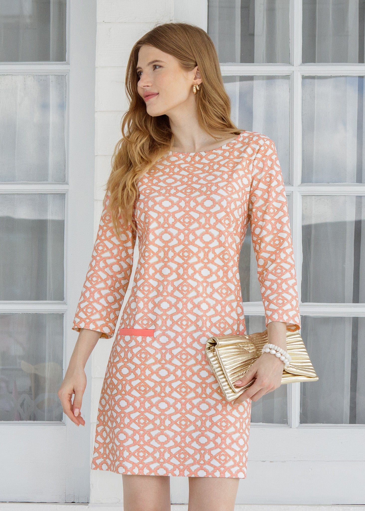 Woman wearing a patterned dress holding a gold clutch in front of a window.