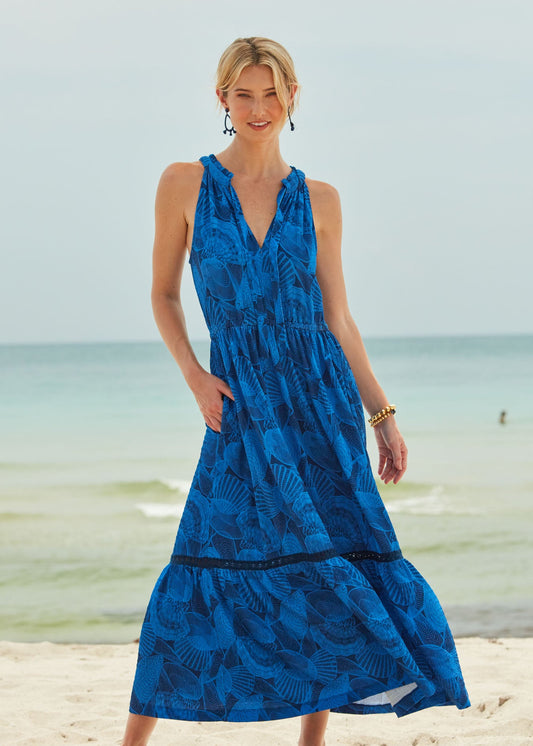 A blonde woman standing on the beach and wearing the flowy San Sebastian Tie Neck Maxi Dress with her hand in the pocket.