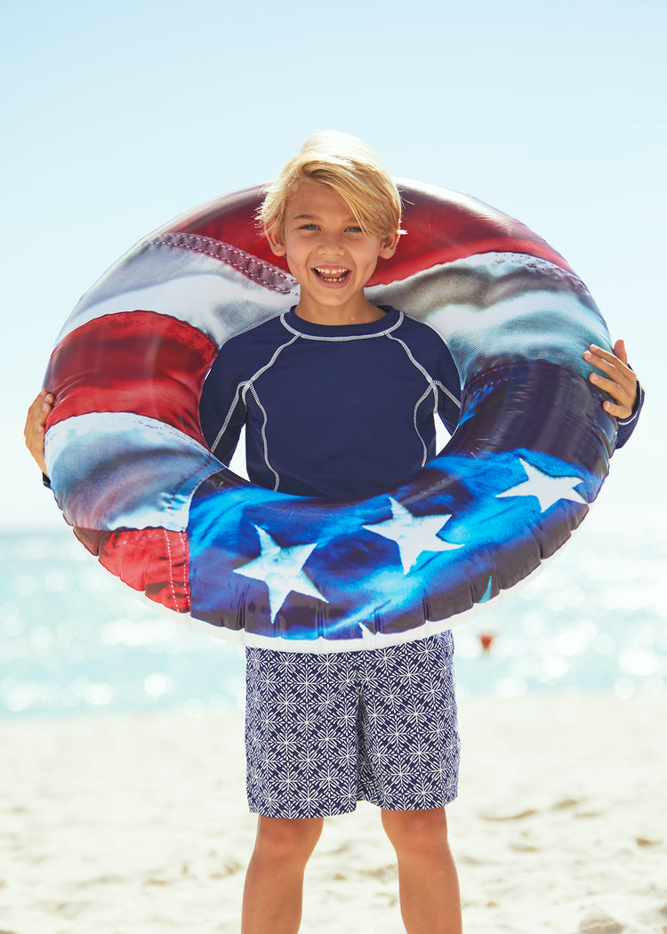 Boy holding inflatable ring on beach wearing Boys Navy Long Sleeve Rashguard with Boys West Indies Swim Trunks