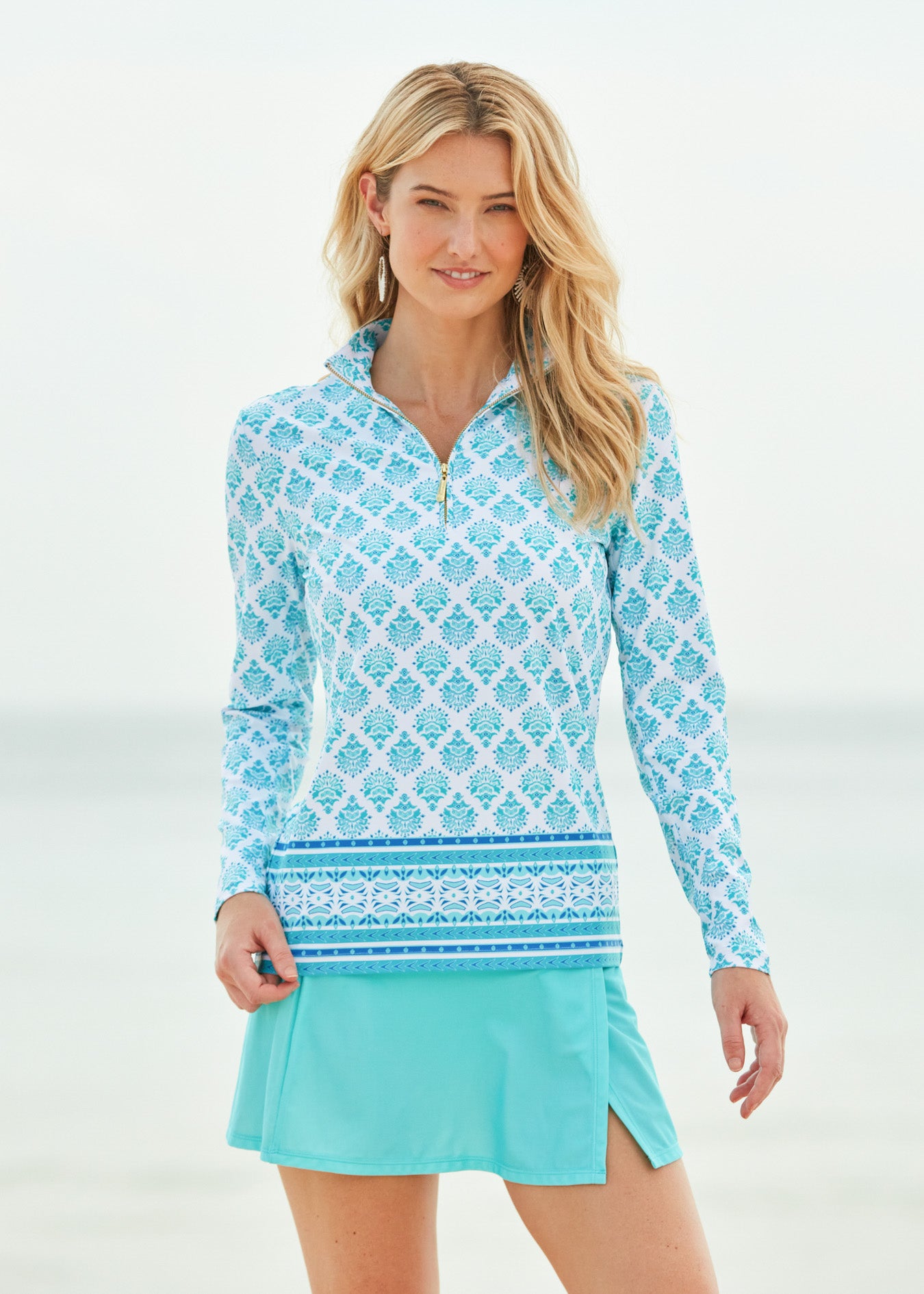 Blonde woman on beach wearing Amalfi Coast Sport Zip Polo with Aqua Classic Swim skirt. 