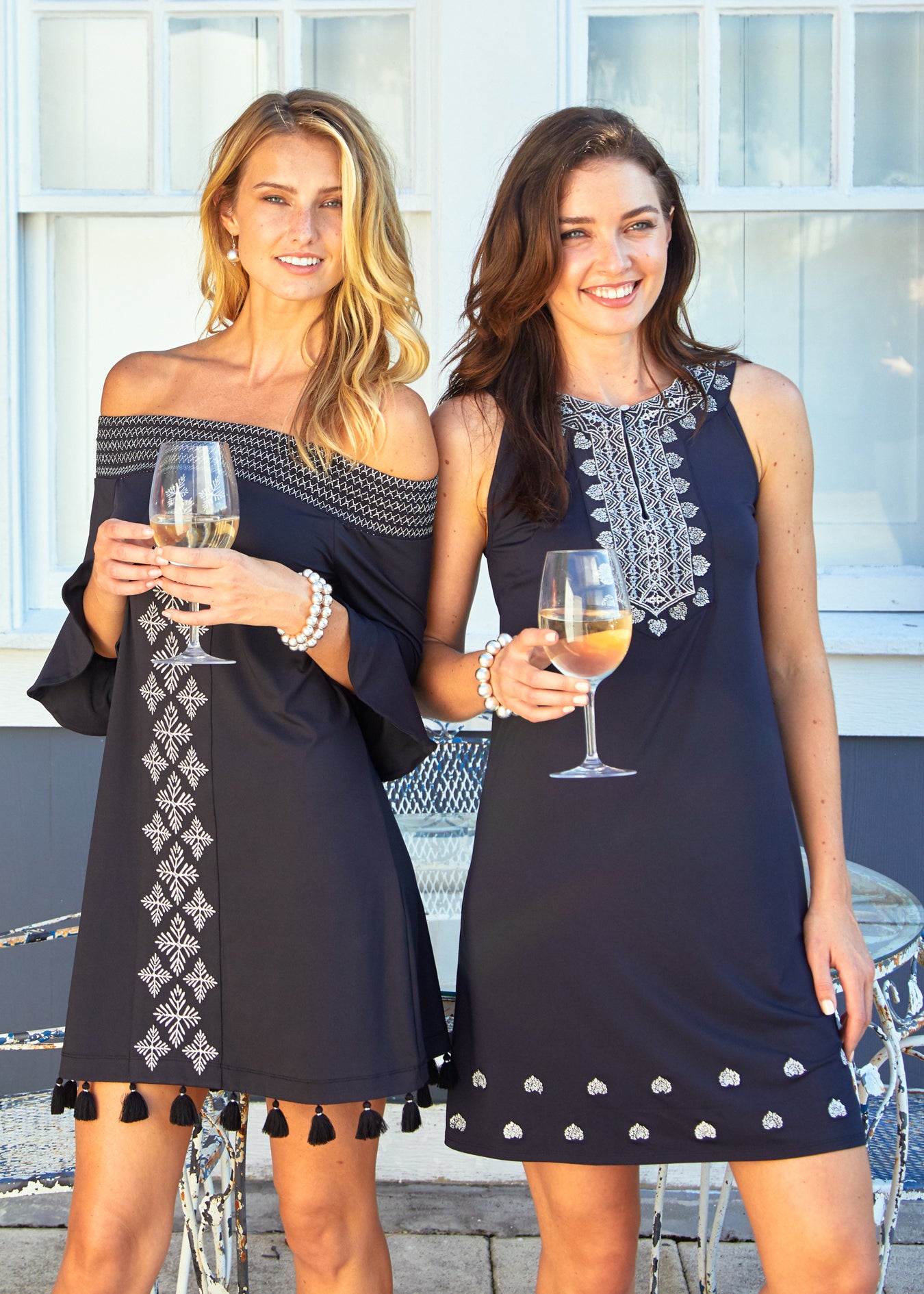 Woman wearing Black Metallic Sleeveless Shift Dress and woman wearing Black/Silver Metallic Off The Shoulder Dress holding white wine