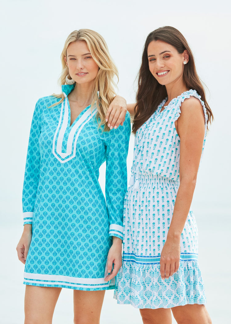 Blonde woman wearing Amalfi Coast Tunic Dress holding drink in hand and with purse on shoulder with Brunette woman wearing Amalfi Coast Smocked Waist Dress on beach