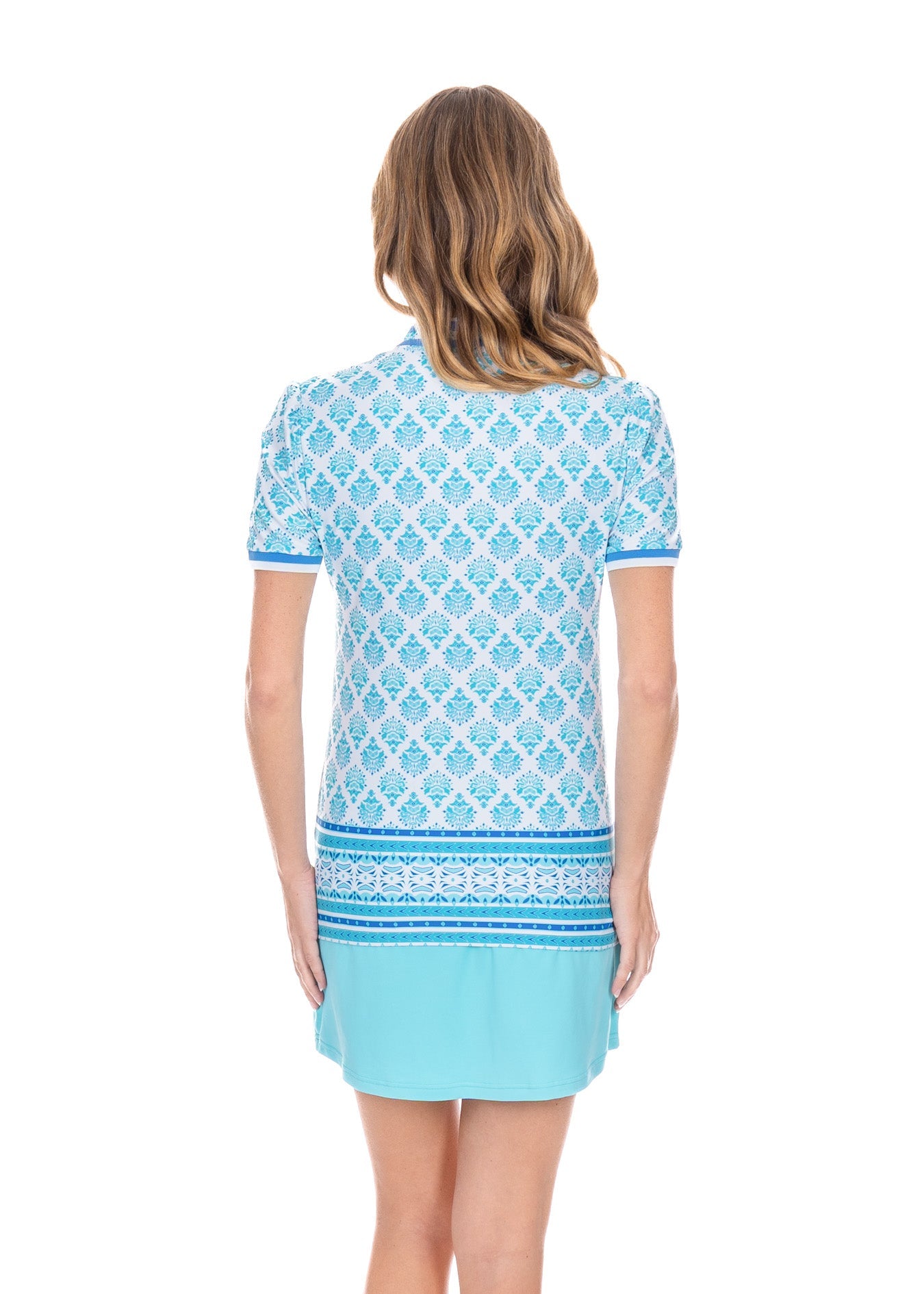 A blonde woman turned around wearing the Aqua Skort and Amalfi Coast Short Sleeve Collared Quarter Zip in front of a white background.