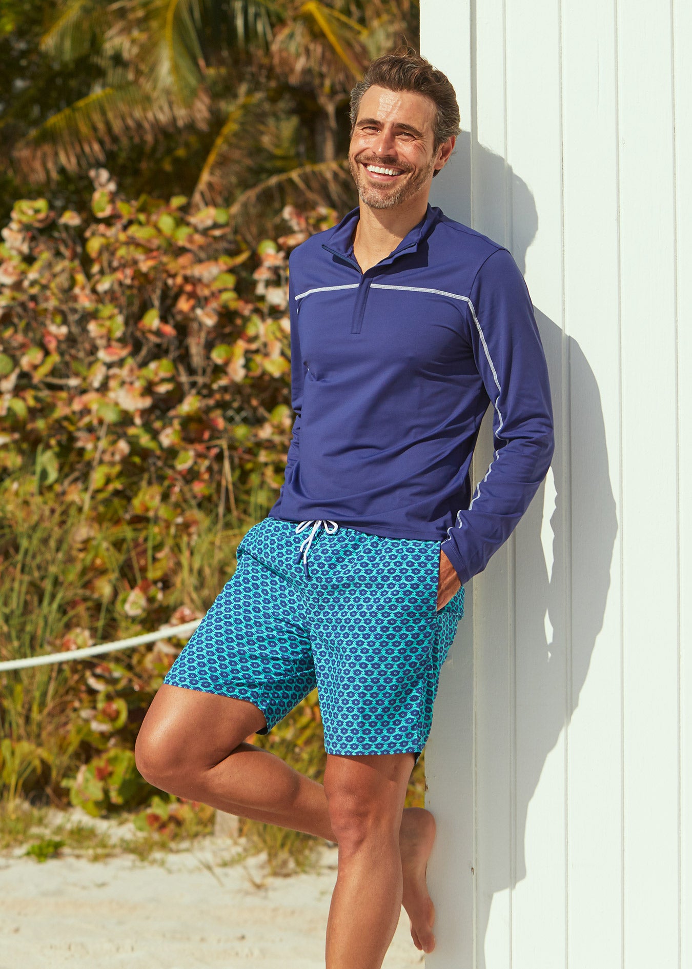 Man leaning against beach shack wearing Cabana Life Navy Sport Zip Top with Aruba Blues Swim Trunks