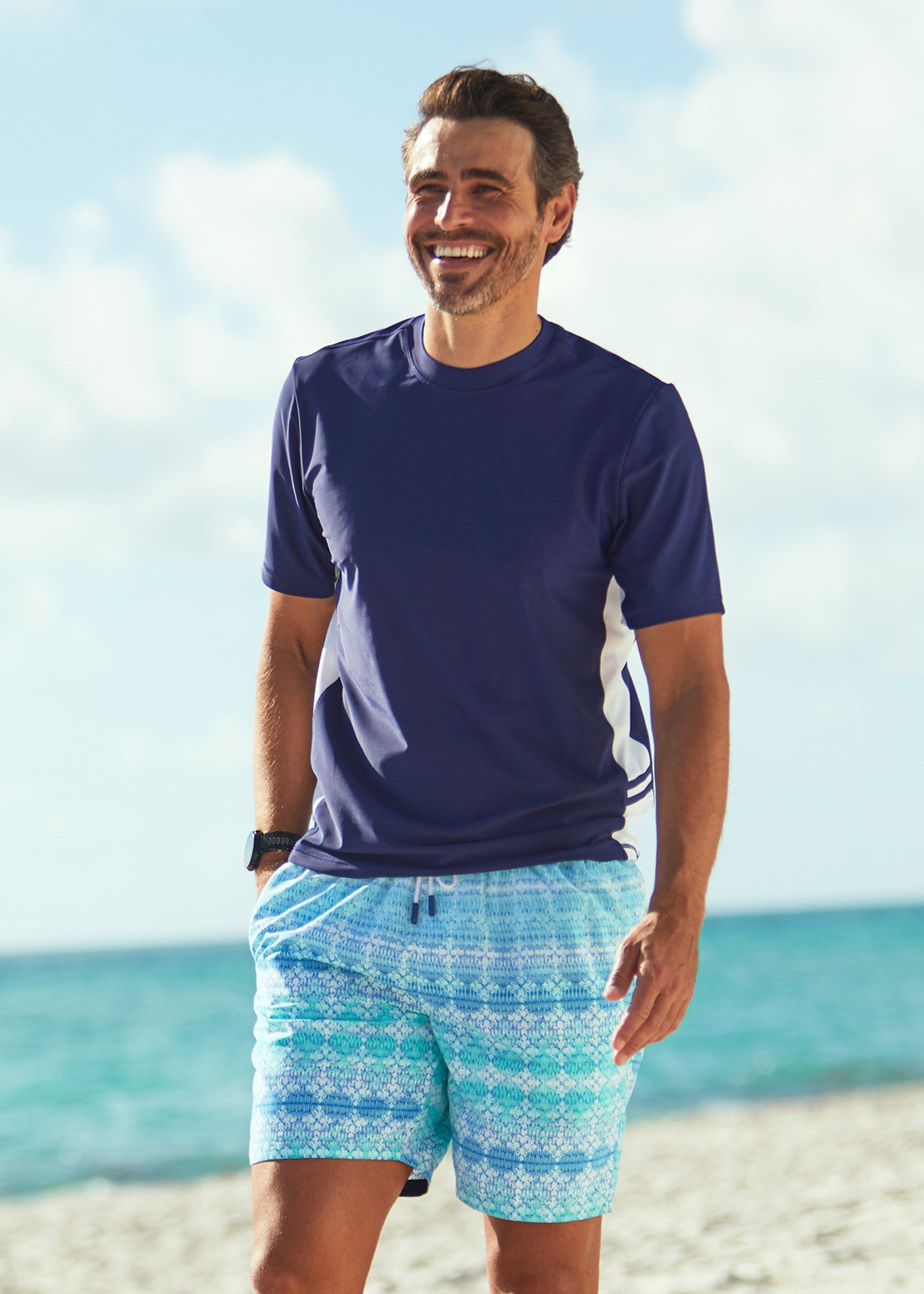 Man walking on beach wearing Cabana Life Navy Short Sleeve Rashguard with Coastal Cottage Swim Trunks